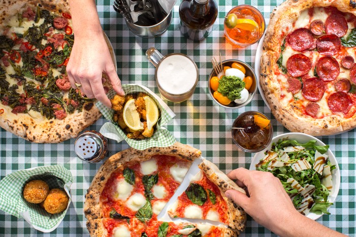 Two different hands reaching across the table for food, with three different Naples-style pizza, salad and drinks