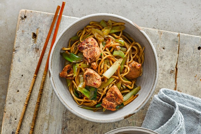 A bowl of chicken chow mein on a grey serving board next to chopsticks