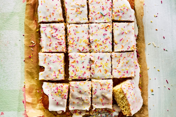 A school cake topped with icing and sprinkles cut into squares on a sheet of brown baking parchment