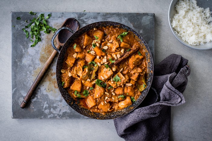 Skillet of vegan jackfruit curry next to a wooden spoon and tea towel