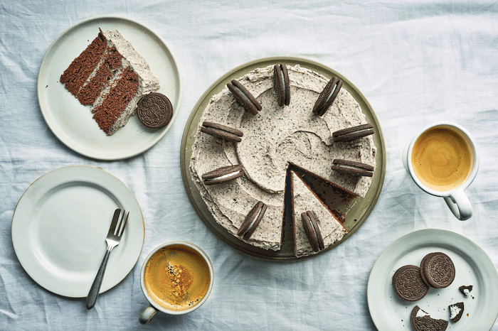 A cake topped with Oreos on a white tablecloth with two cups of coffee